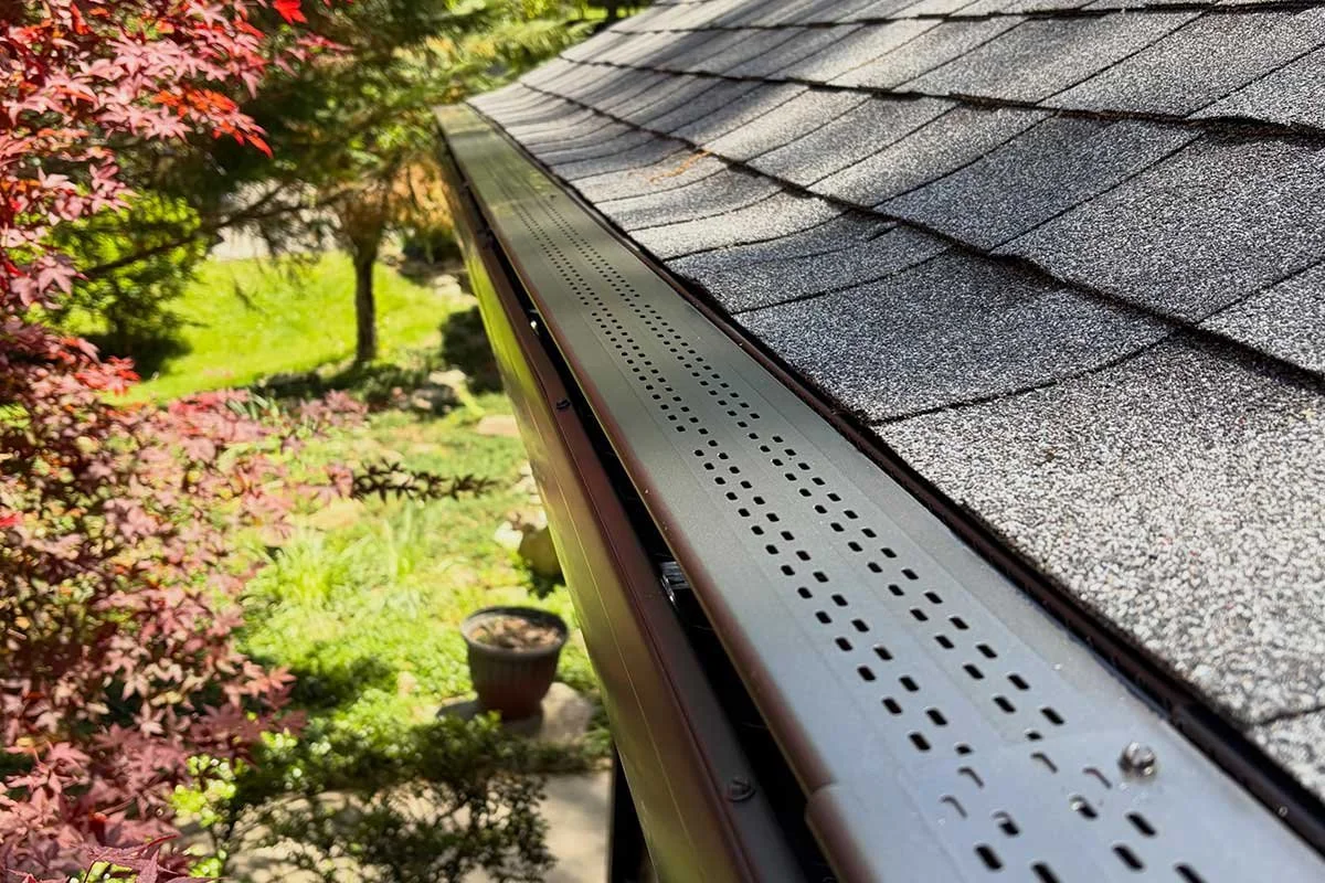 Close-up of a house's roof showing a metal gutter and gray asphalt shingles with trees and a garden in the background.