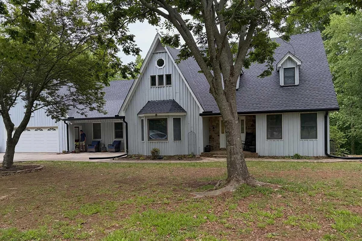 A light gray house with a pitched roof, multiple windows, and a small front porch surrounded by trees and a grassy yard.