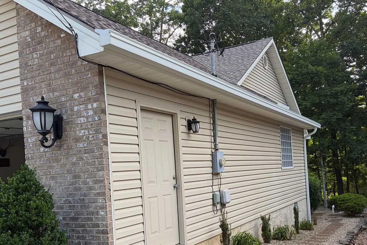 Side of a house with beige vinyl siding, a brick section, and outdoor lighting fixtures, surrounded by small bushes and trees.
