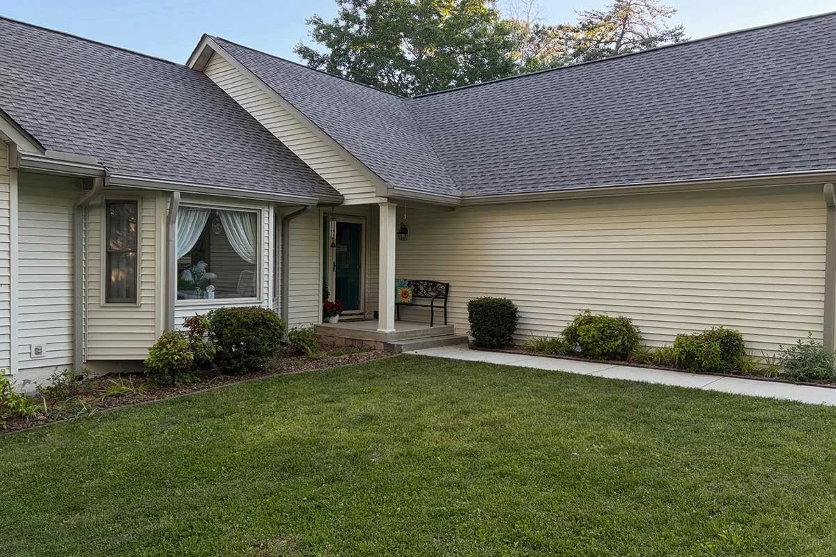 Front view of a house with a well-maintained lawn, beige siding, and a gabled roof. There is a small porch with steps and a door, a bench, and some bushes along the house.