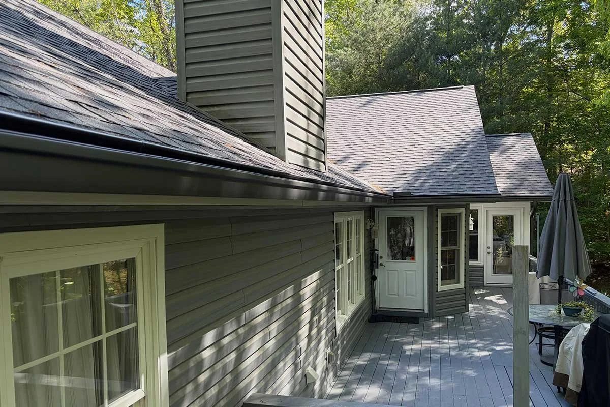 A wooden deck attached to a house with gray siding, white-trimmed windows, a white door, and an outdoor table with an umbrella, surrounded by green trees in the background.