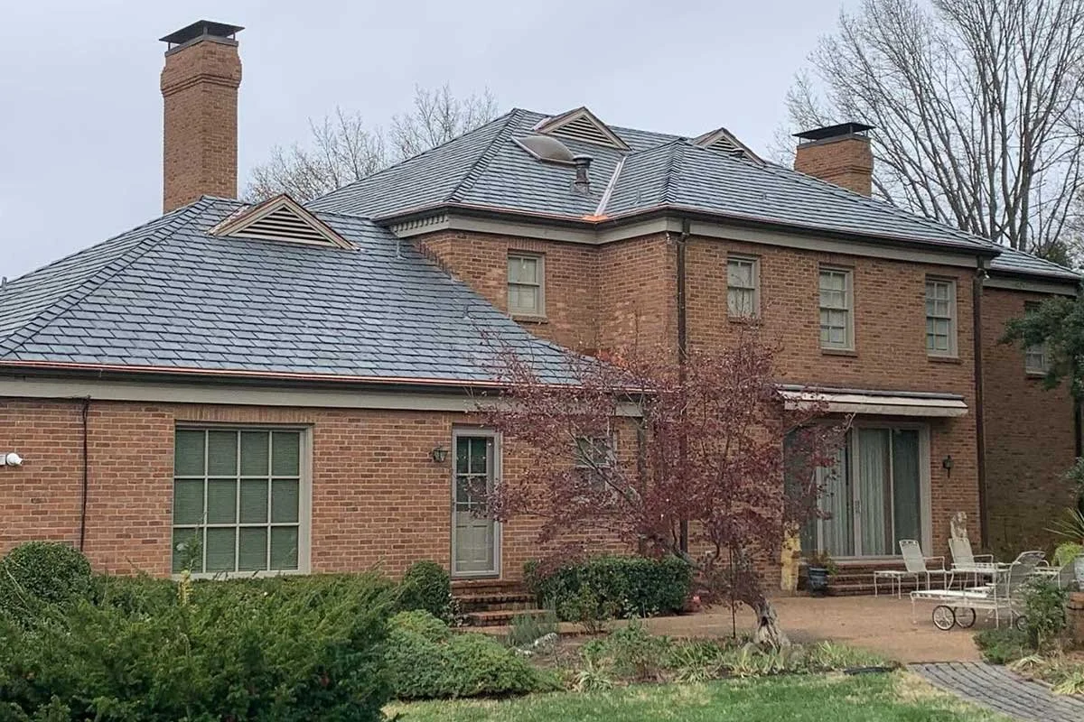 Rear view of a brick house with a tiled roof, chimneys, and a backyard patio with outdoor furniture.