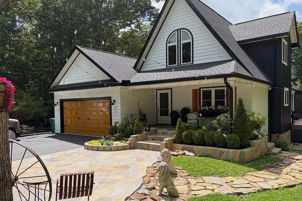 Front view of a modern house with white siding, black accents, and a gabled roof, featuring a landscaped front yard with stone pathways, shrubs, and a decorative statue of a horse.