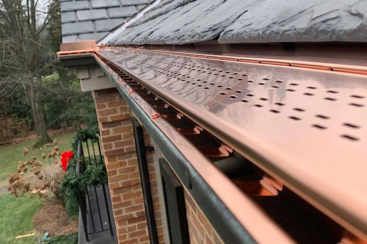Close-up view of a special-order copper LeafX gutter protection system attached to the roof of a brick house, with trees and a garden in the background.