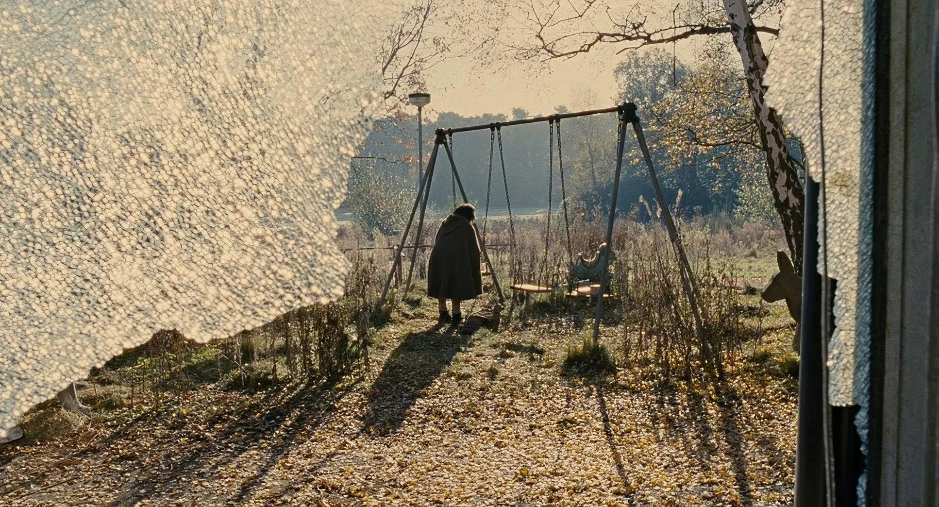 Kee sits on a swing, seen through the shattered window of an abandoned playground in Children of Men.