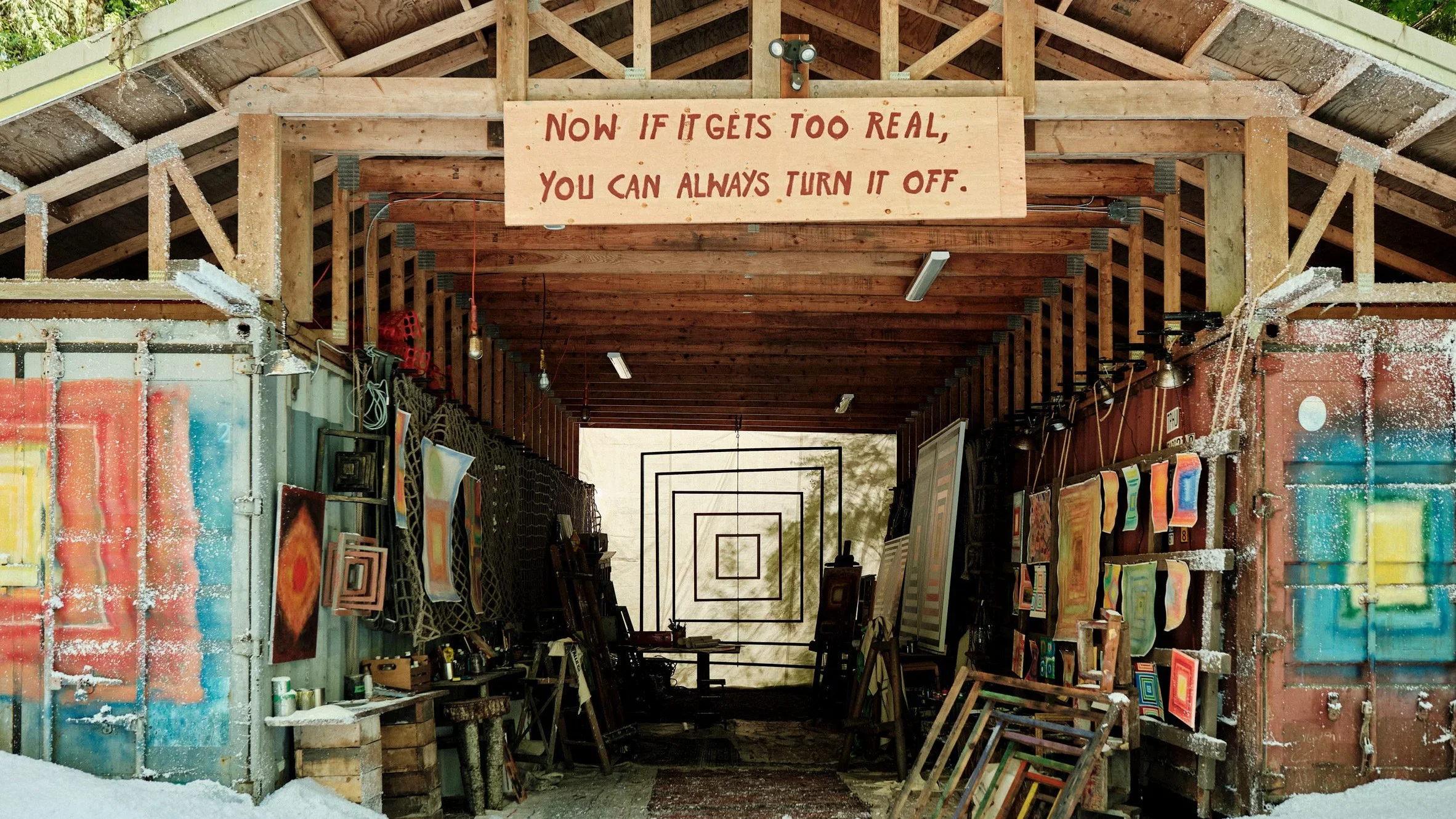 An open wooden studio filled with paintings, canvases and art supplies. A sign overhead reads: "Now if it gets too real, you can always turn it off."