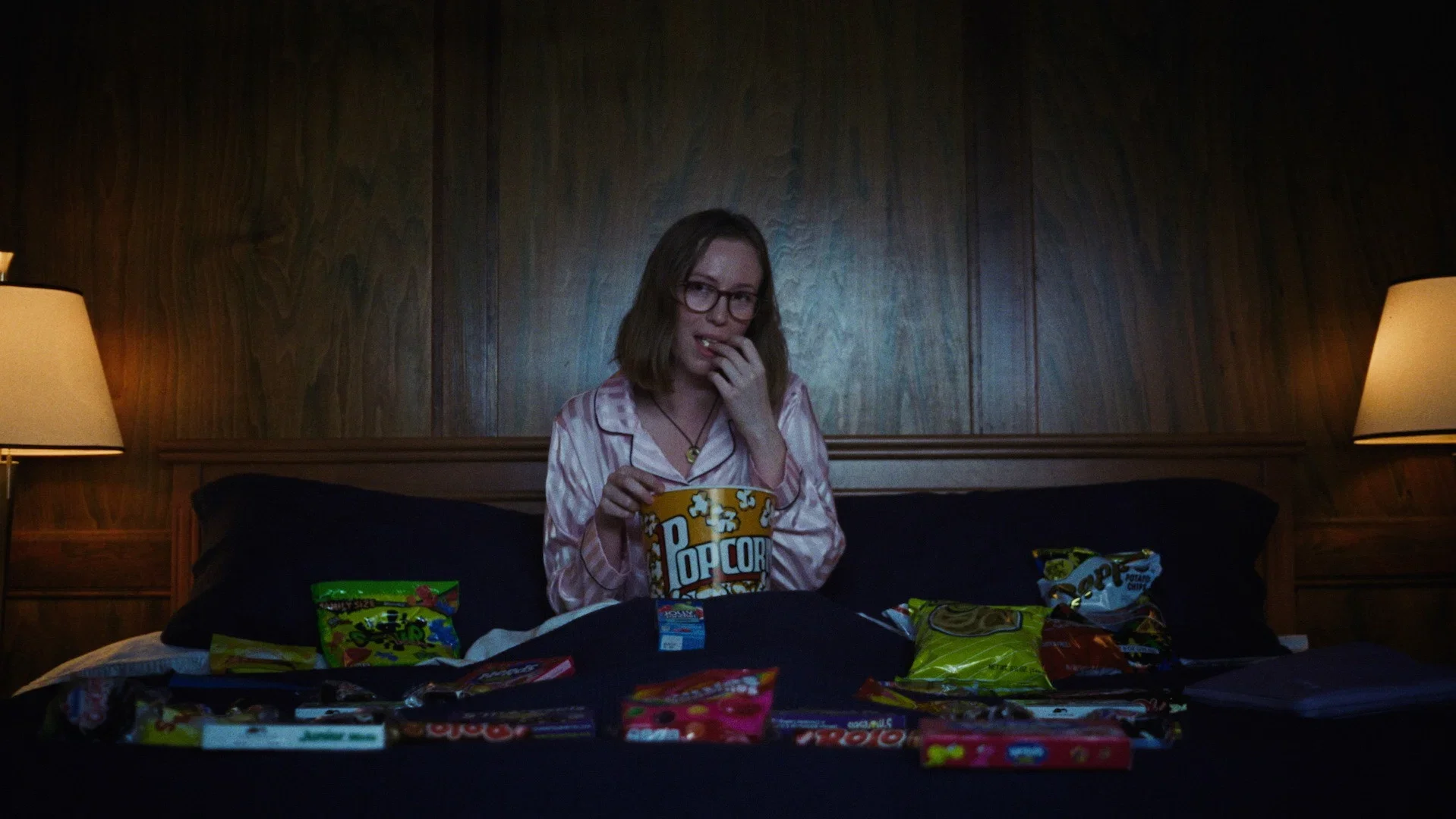 A woman wearing glasses and pink striped pyjamas sits on a bed eating popcorn, surrounded by an assortment of sweets, crisps and other cinema snacks.