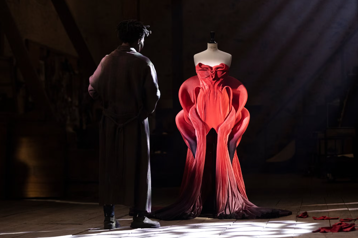 Sam stands beside a sculptural red gown on a mannequin, illuminated by a shaft of light in a dark workshop.