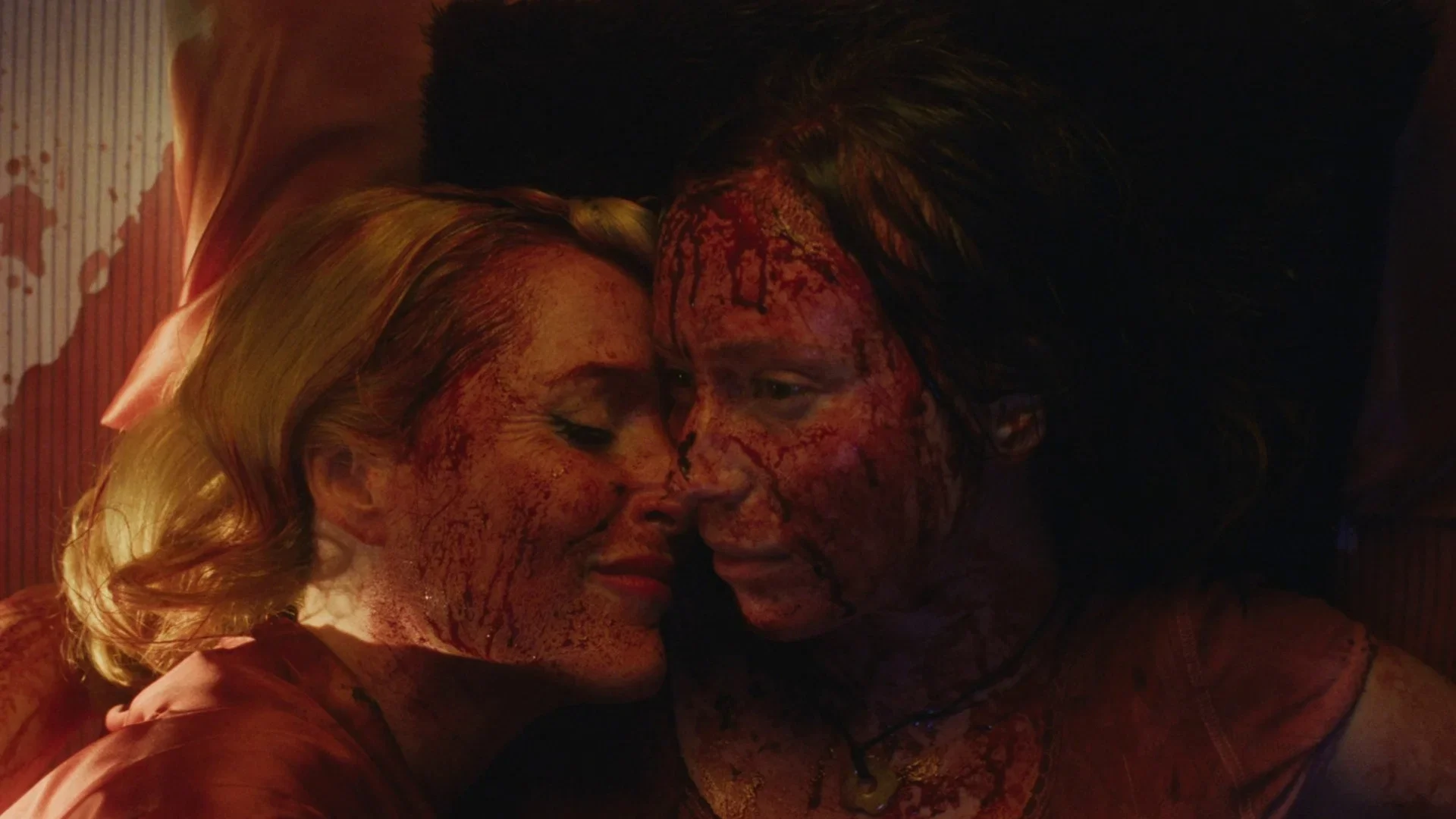 Two women, their faces and clothing covered in blood, lie cheek to cheek in an intimate embrace, illuminated by warm red light.