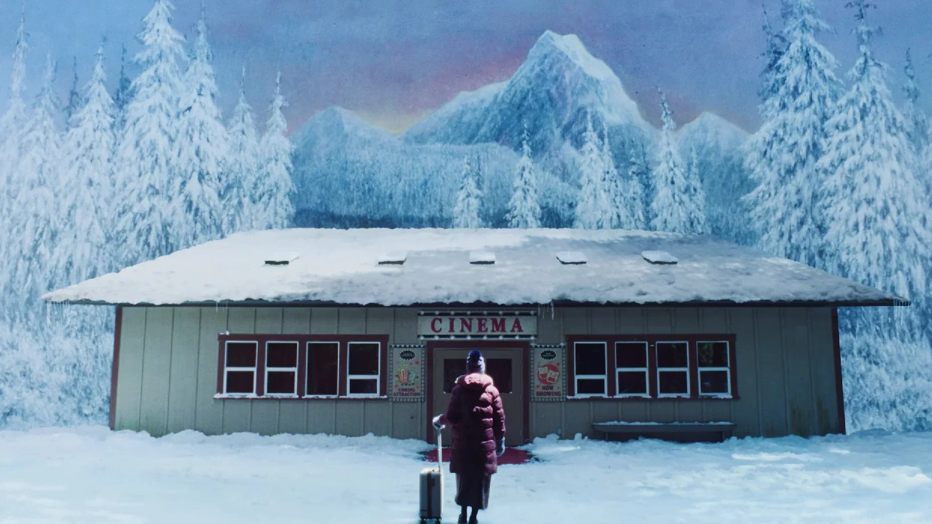 A solitary figure with a suitcase stands outside a small cinema surrounded by snow-covered trees and a painted mountain landscape.