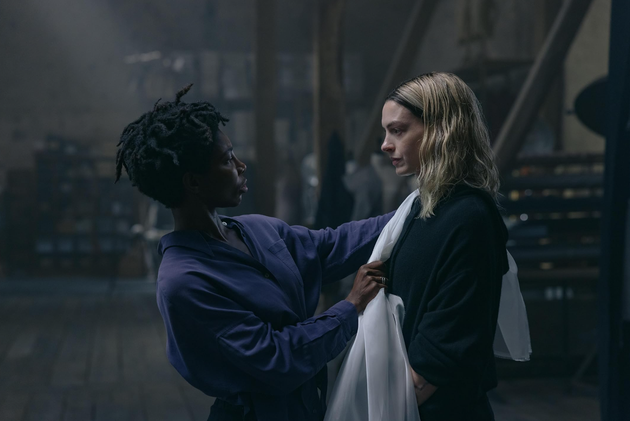 Sam adjusts white fabric around Mary's shoulders during a fitting in their dimly lit workshop.