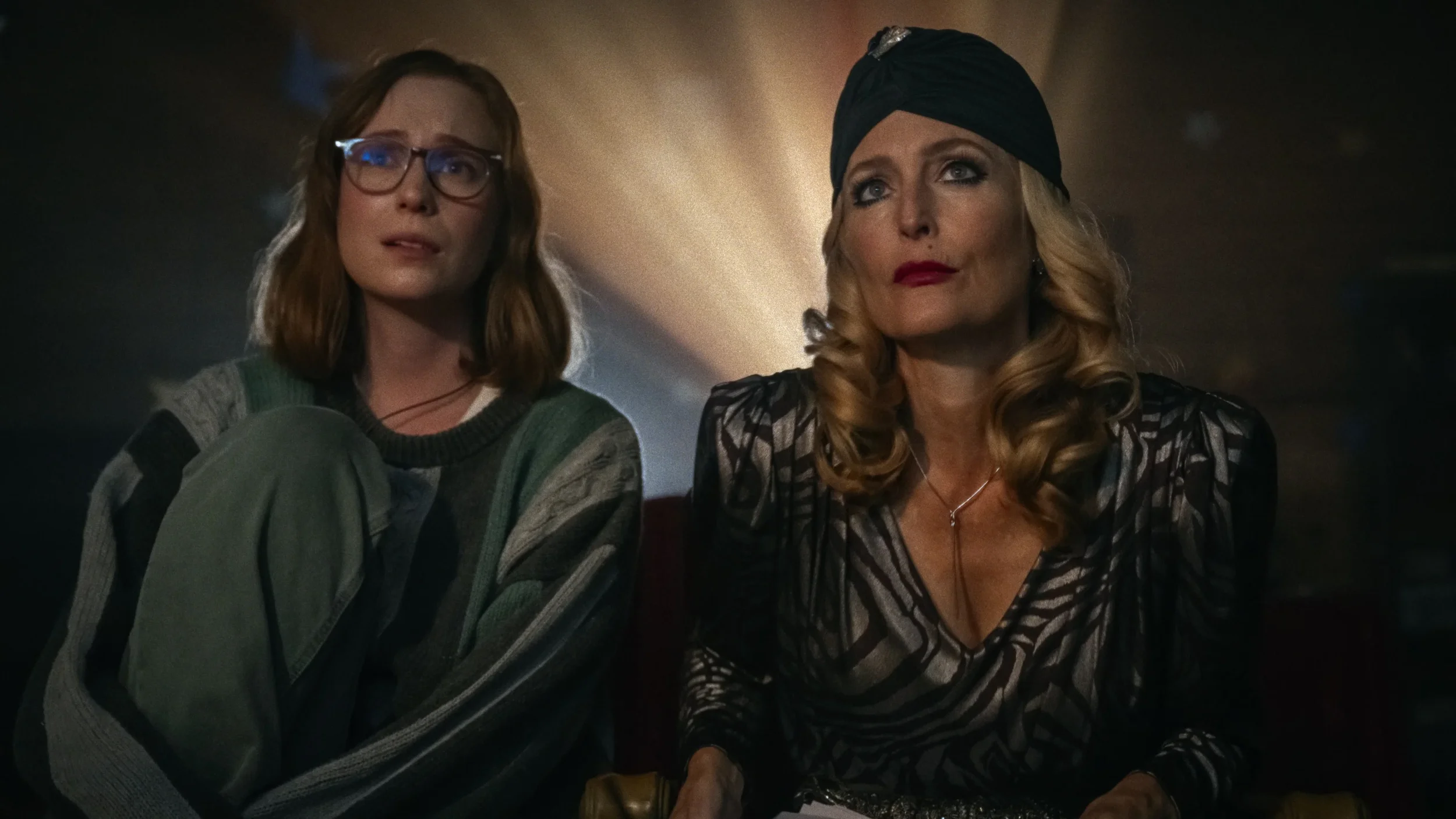 Two women sit side by side in a dimly lit room, one wearing glasses and a green jumper, the other with blonde curls, a black headscarf and patterned clothing.