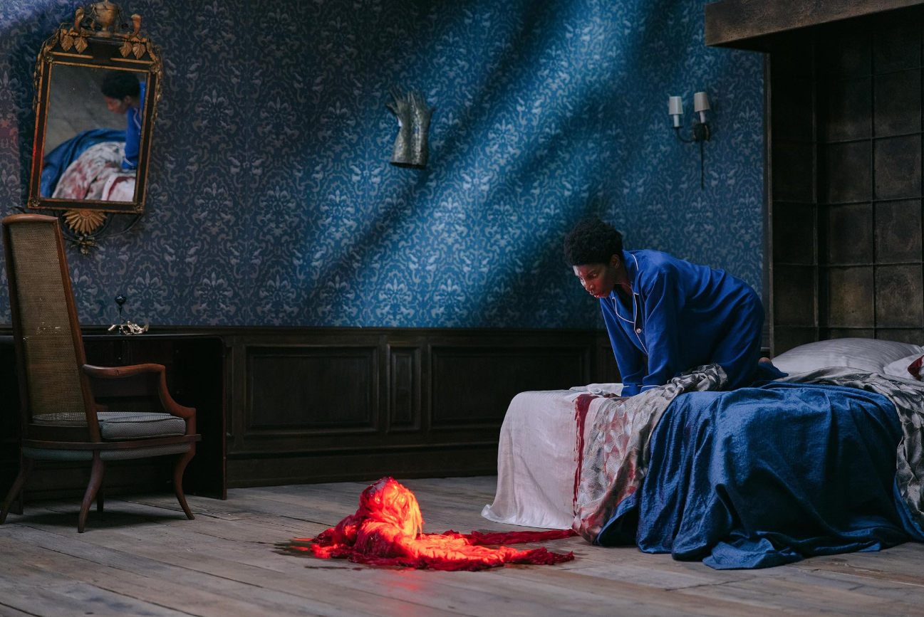 Sam leans over a bloodstained bed as a glowing red fabric apparition lies on the wooden floor.