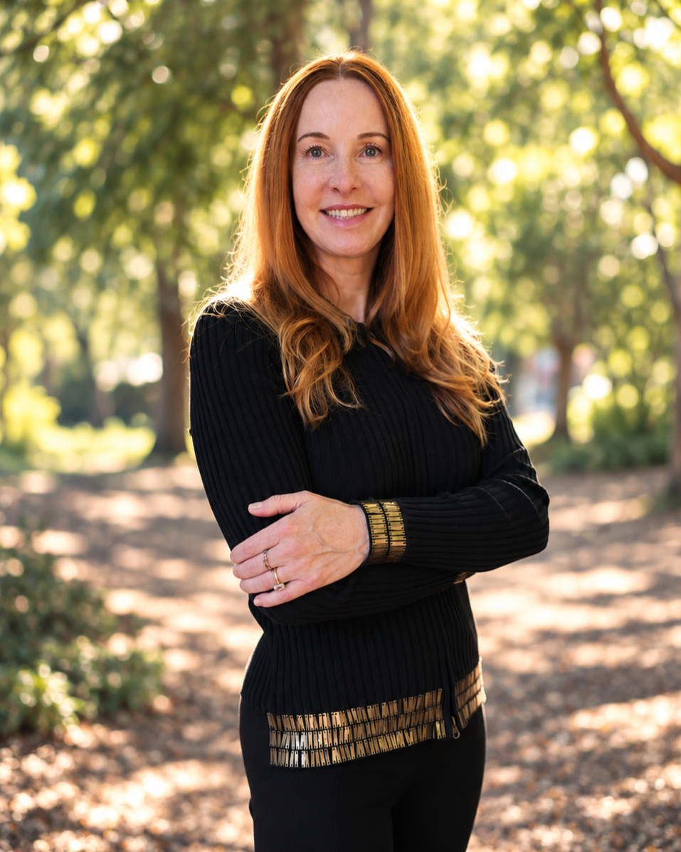 A woman with long red hair is smiling outdoors, standing with arms crossed in a park with trees and sunlight in the background.