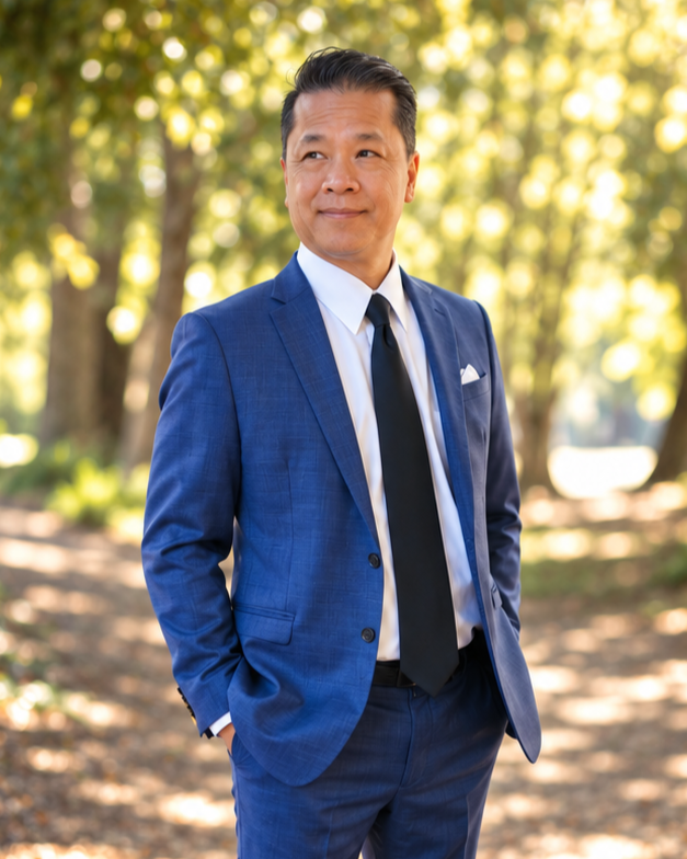 A man in a blue suit and black tie standing outdoors in a park with sunlight filtering through trees.