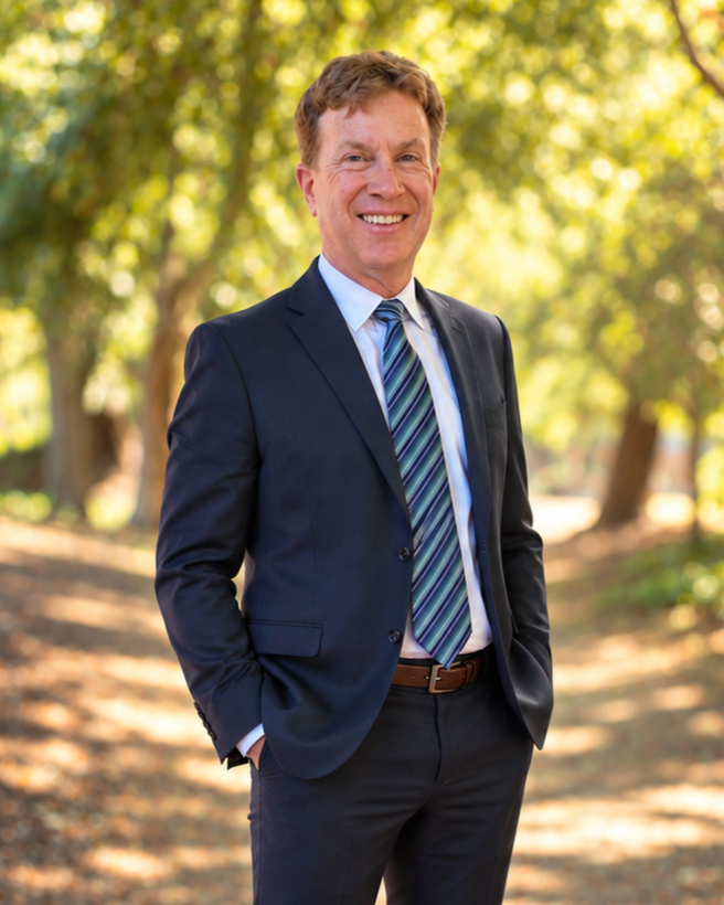 Smiling man in a suit standing outdoors in a park with trees and sunlight