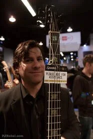 Person holding an electric guitar at a trade show or exhibition with others in the background.