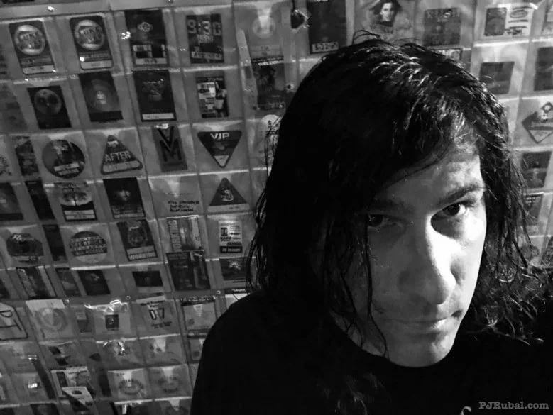 Young person with wet, wavy hair taking a black and white selfie with a wall of posters in the background.