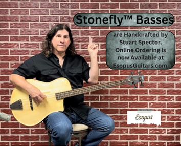 A man with long dark hair sitting on a chair, holding an acoustic guitar, in front of a brick wall with a sign promoting Stonefly™ Basses and online ordering from Esopus Guitars.