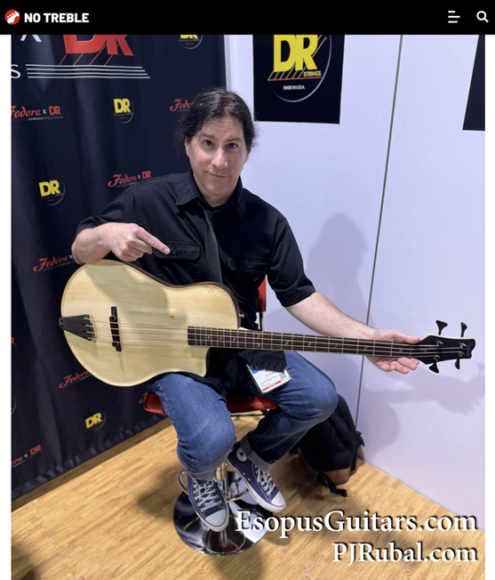 A person sitting on a stool holds an acoustic guitar and points at it while looking at the camera. The background features logos and signs related to guitar strings and a website address.