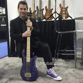 Man sitting on the floor with a purple electric guitar, smiling, at a music exhibition with guitars displayed behind him.