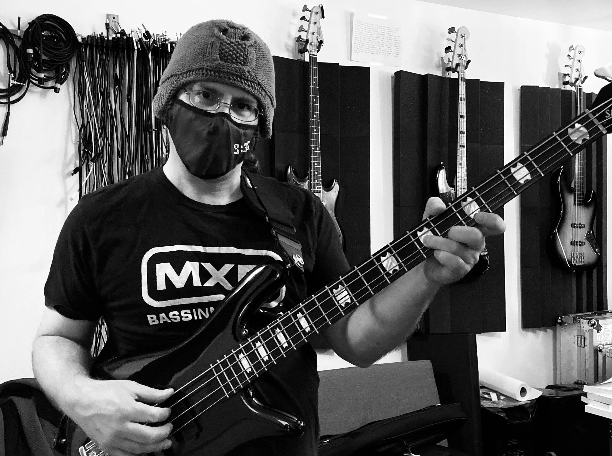 A person wearing glasses, a face mask, and a knitted owl hat is holding an electric bass guitar. He is standing in a music studio with guitars hanging on the wall behind them and various musical equipment visible.