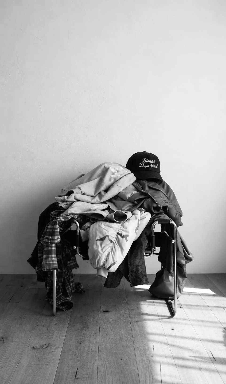 A pile of clothes on a metal chair with a black cap reading 'Blondes Day Ahead' on top, against a plain wall and wooden floor.