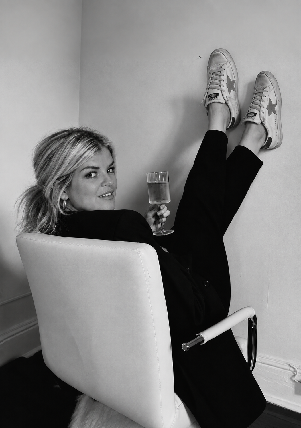A woman sitting in a chair with her legs up on the wall, holding a glass of wine and looking at the camera, wearing Converse sneakers and a black outfit, in a black and white photo.