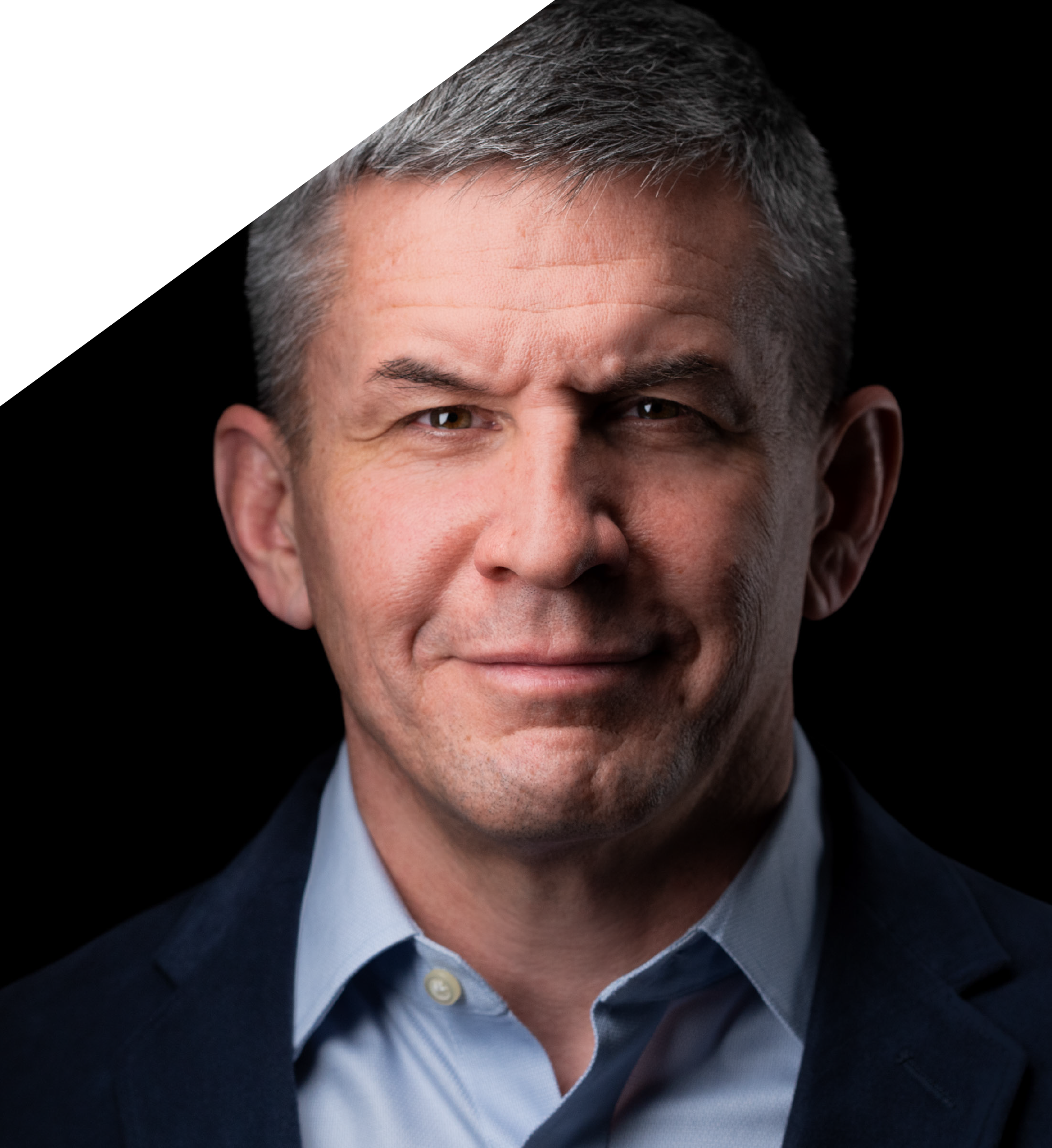 Close-up of a middle-aged man's face and upper torso, with short gray hair, brown eyes, wearing a dark suit and a light blue shirt, against a black background.