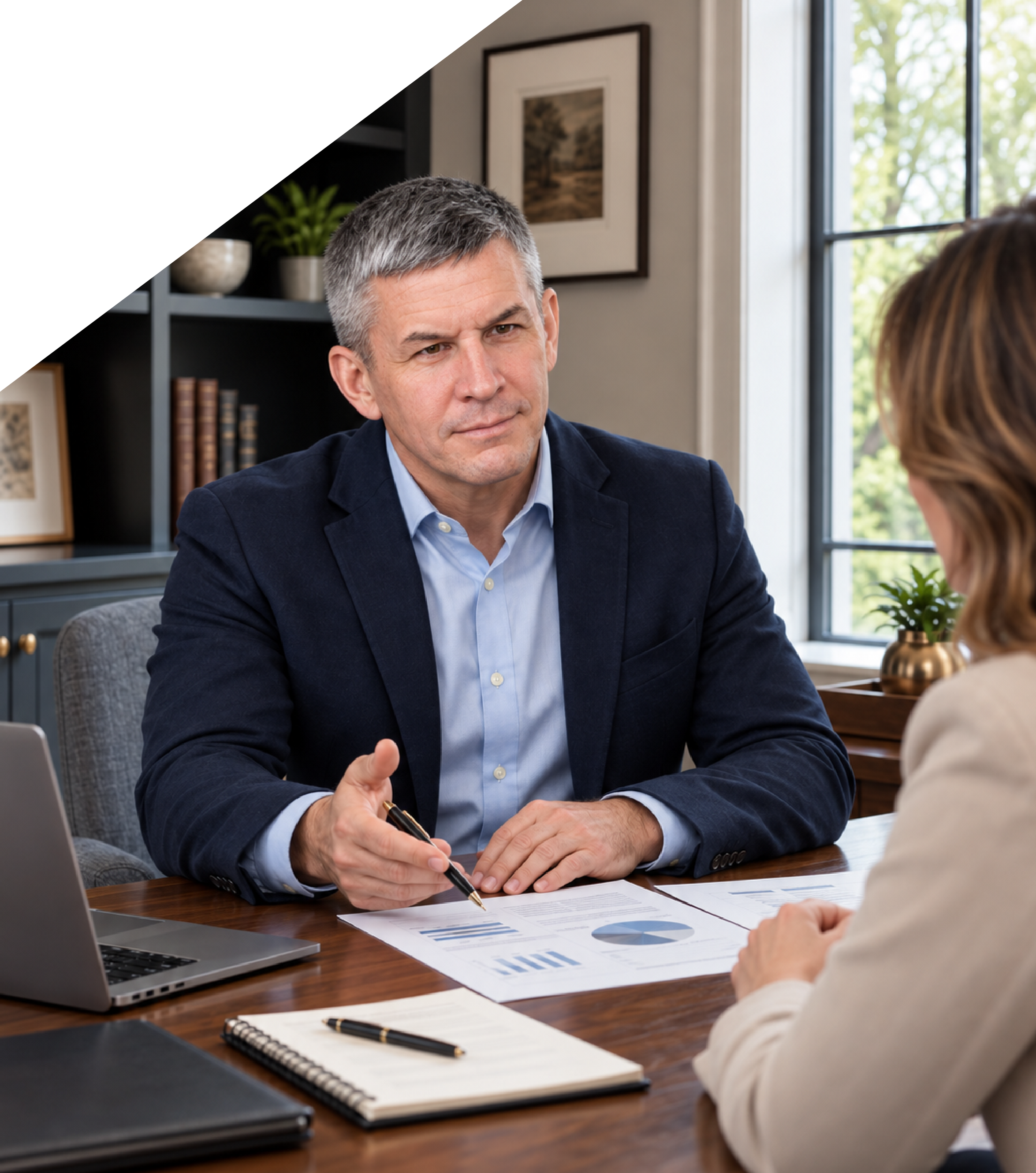 A middle-aged man in a navy blazer and light blue shirt gesturing and holding a pen while sitting at a wooden desk during a meeting with a woman. The desk has documents with graphs, a notebook, folded papers, a laptop, and pens. The background features a window with greenery outside, a black bookshelf with books and framed photos, and a framed picture on the wall.