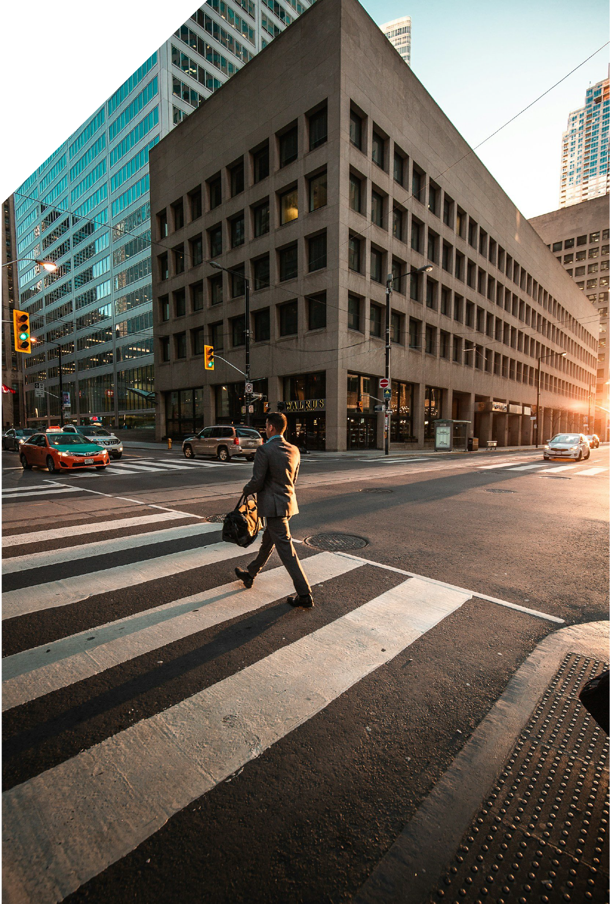 A man in a business suit crossing a city street at a crosswalk during sunset, with tall office buildings on both sides and traffic lights.