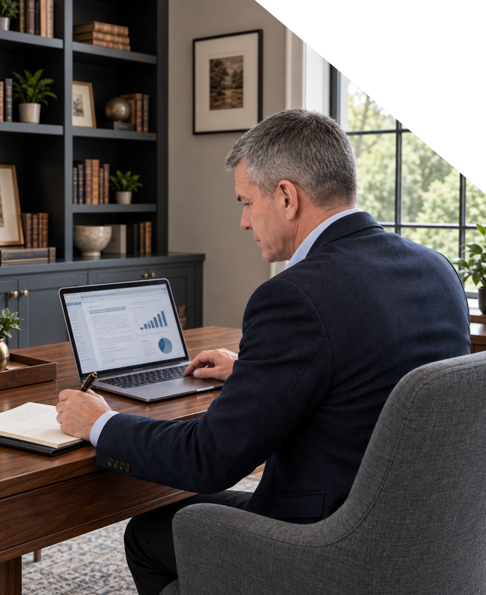 A middle-aged man with gray hair in a suit sits at a wooden desk in a well-lit room, looking at a laptop displaying charts and graphs. He has a pen in his right hand and an open notebook on the desk. There are bookshelves with books and decorative items behind him, and a window letting in natural light.