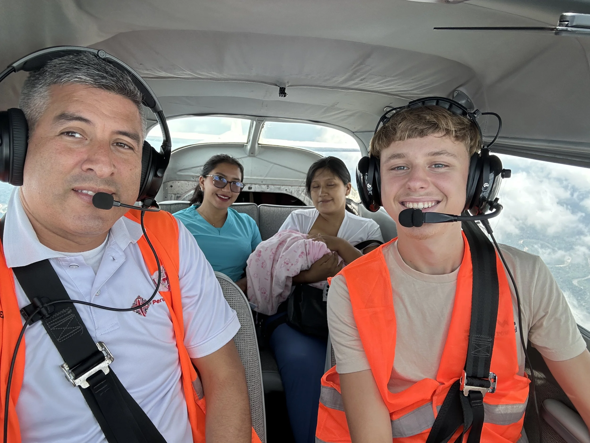 Four people sitting inside an airplane while one of them holds an infant. The two pilots are wearing orange safety vests and headsets, looking at the camera and smiling.
