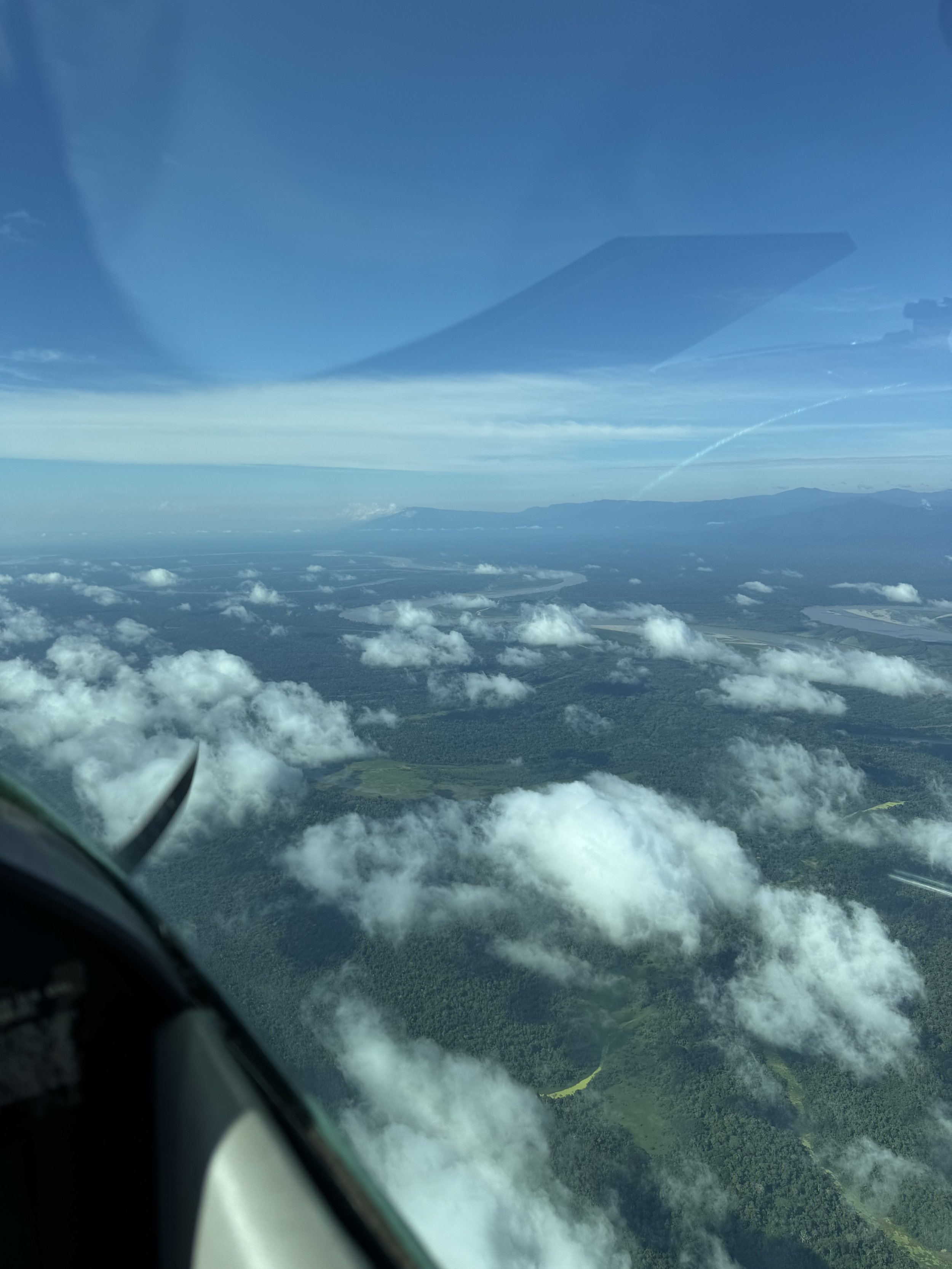 Aerial view from an airplane showing clouds, a river, green land, and distant mountains under a blue sky.
