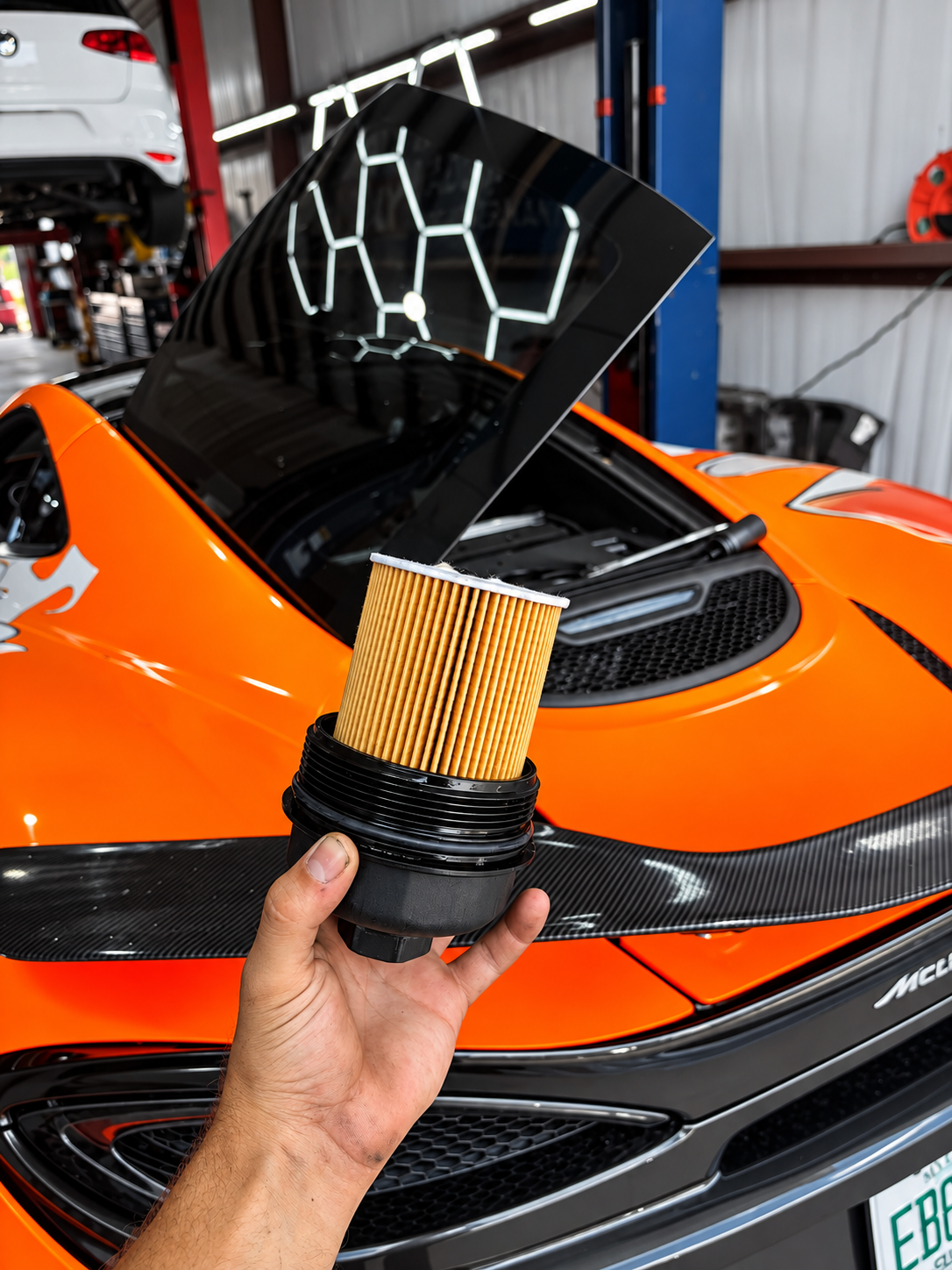 Person holding a car air filter in front of an orange sports car inside a garage or workshop.