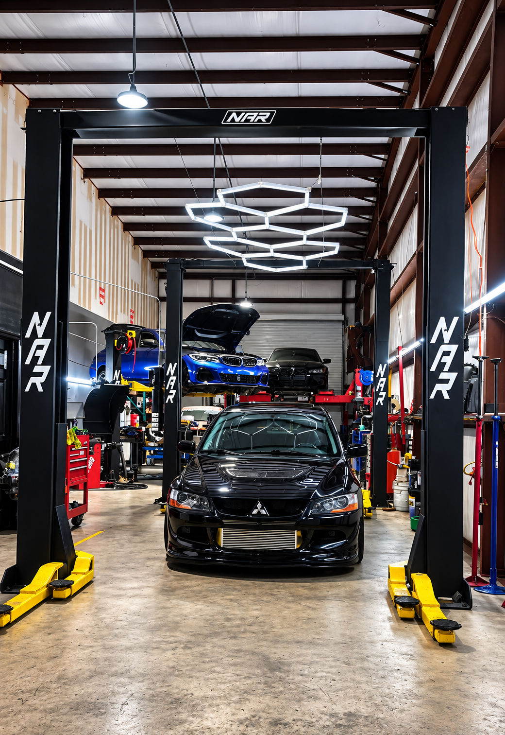 Inside an auto repair shop with a black Mitsubishi Lancer Evolution in the foreground, two cars on lifts in the background, car repair equipment, tools, and bright lighting.