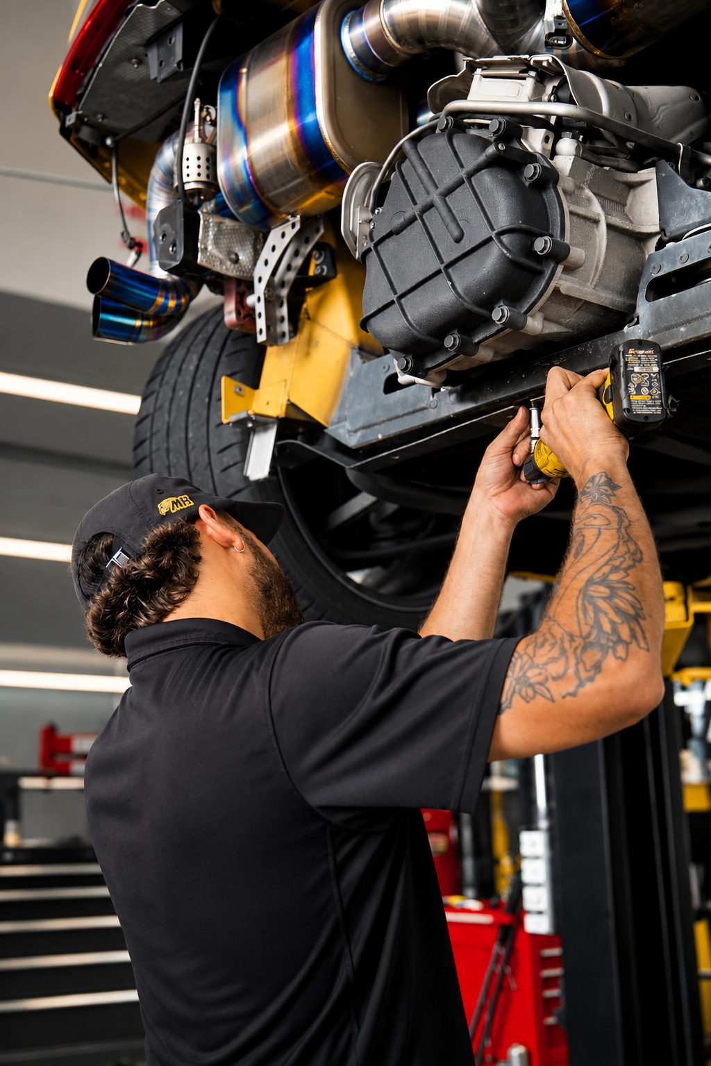 A mechanic working on the underside of a race car, using a power drill, with a focus on the car's exhaust system and engine components.