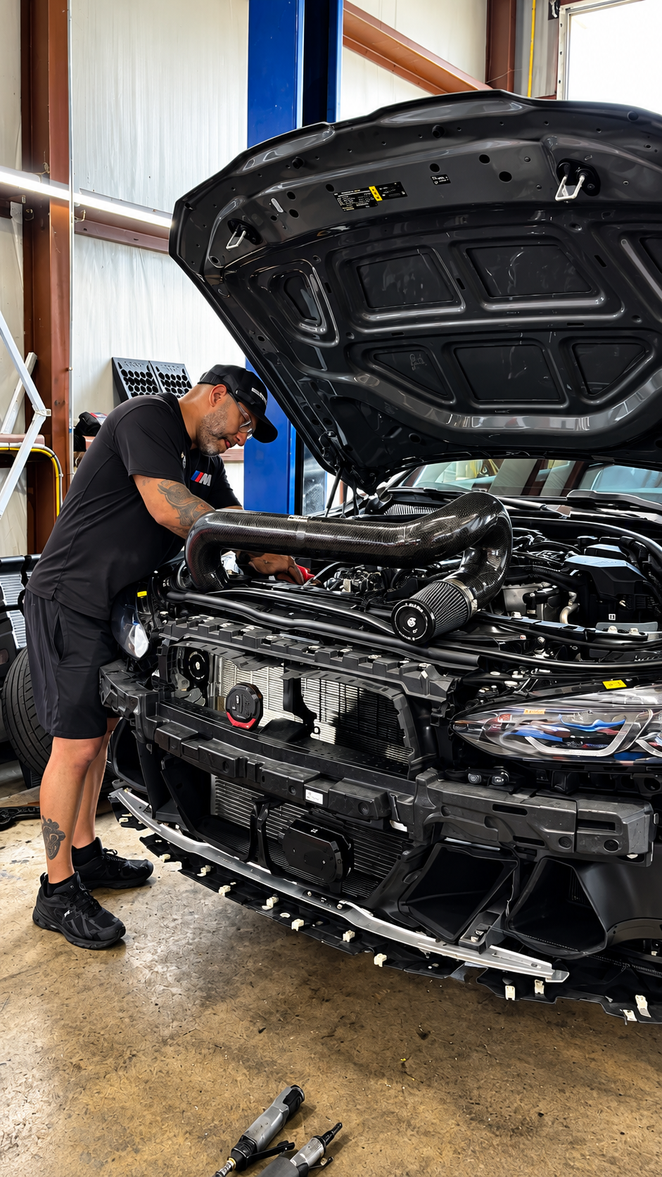 A man with tattoos working on a car engine inside a garage, with the car's hood open, exposing high-performance parts and a large carbon fiber air intake pipe.