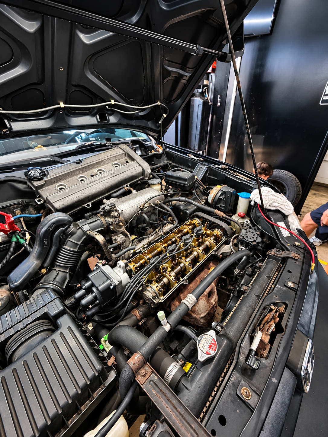 Open car hood showing engine with visible components including the valve cover, spark plug wires, and oil cap, in a workshop.