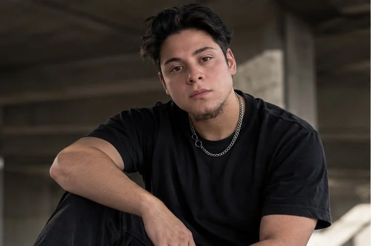 Young man with dark hair, wearing a black T-shirt and silver chain necklace, sitting with arm rested on his knee in an industrial setting.