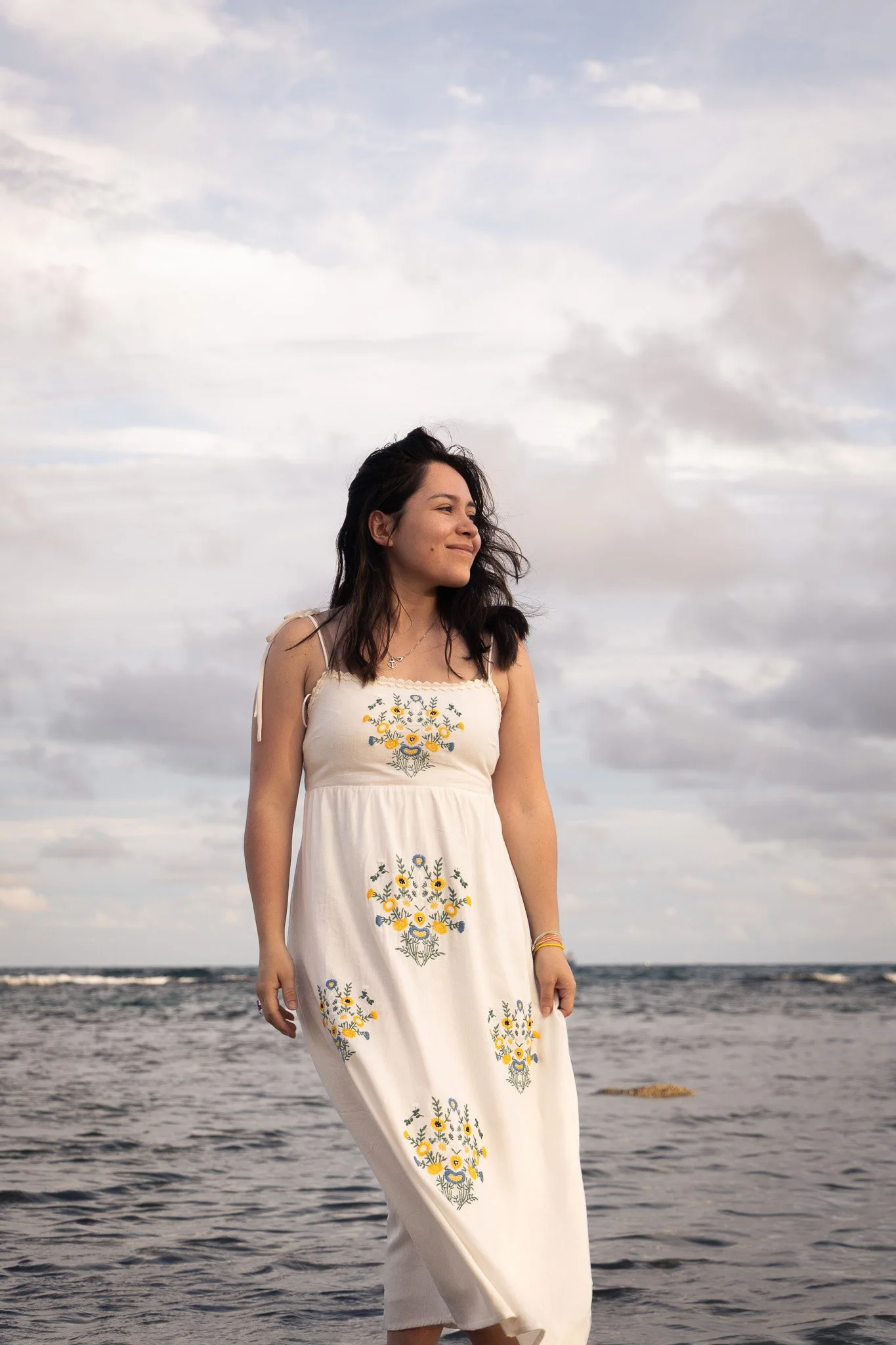 A woman with dark hair standing on a beach with water and cloudy sky in the background, wearing a white dress with floral embroidery.