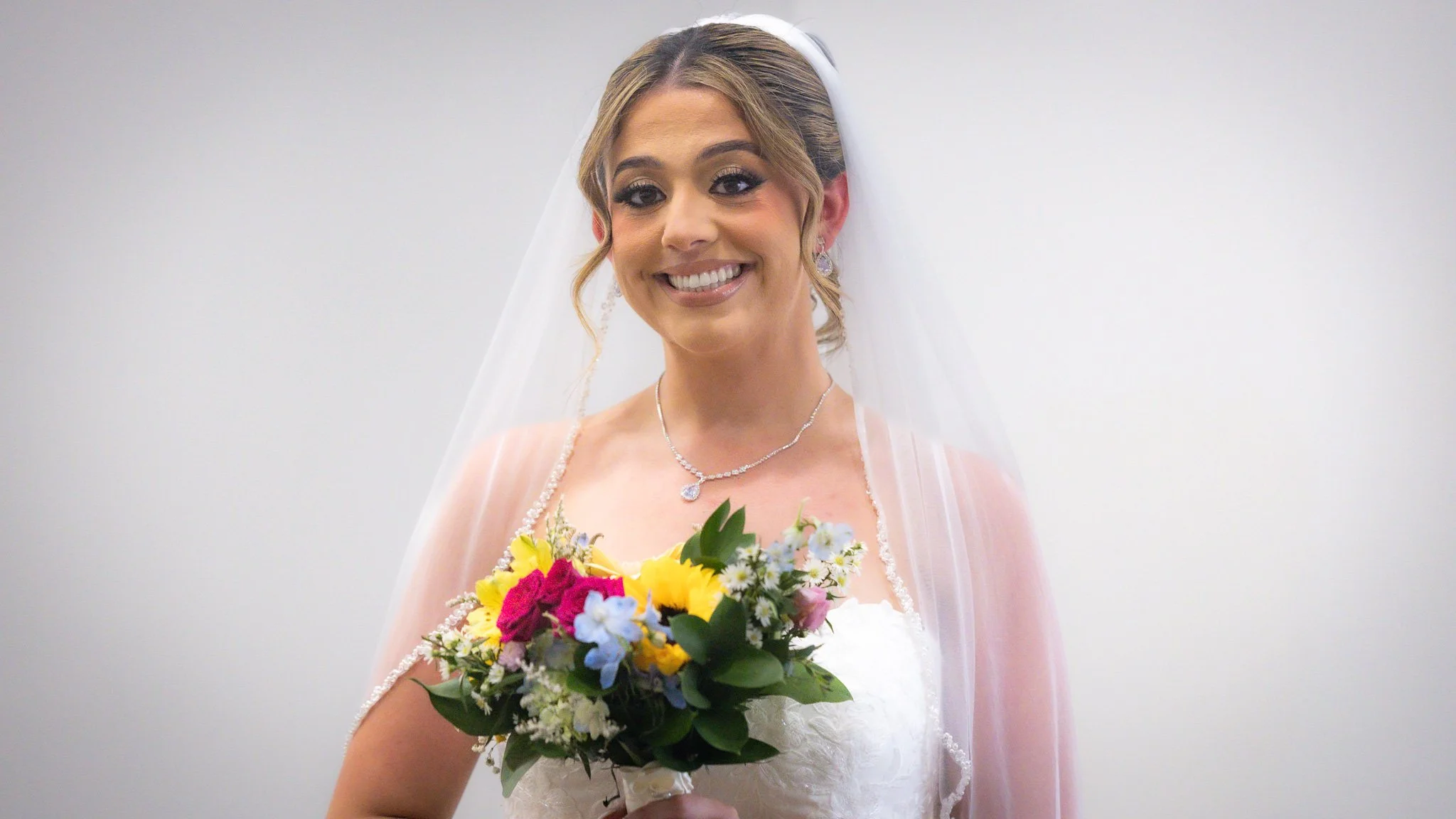 A smiling bride with light brown hair styled in an updo, wearing a white wedding dress and a veil, holding a colorful bouquet of flowers.