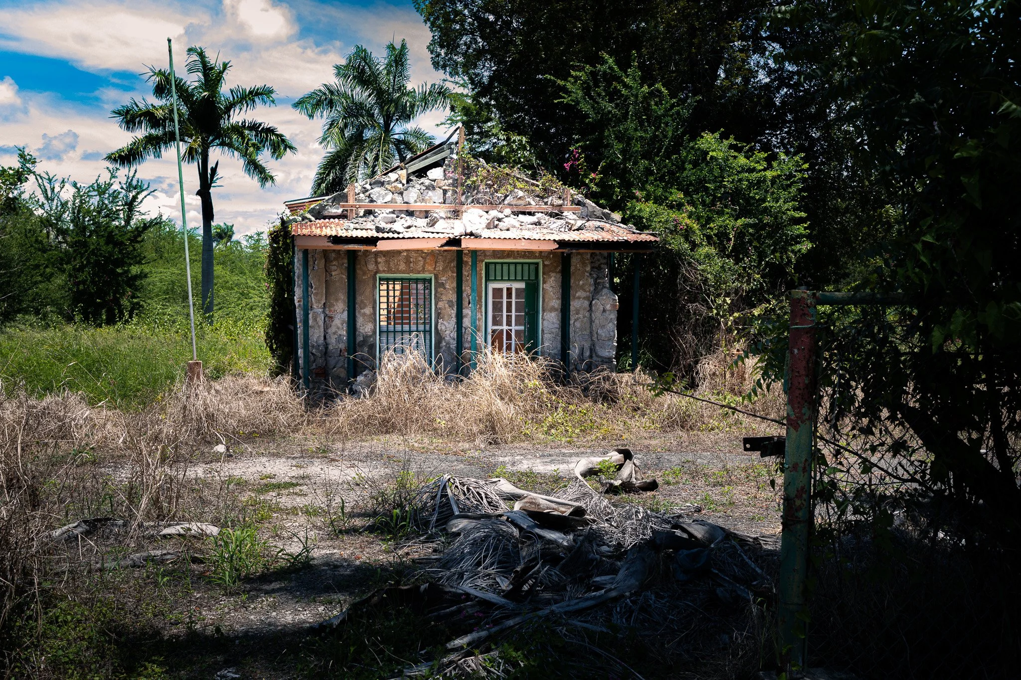 A partially destroyed stone house with a damaged roof, surrounded by overgrown grass, trees, and bushes, with a sky filled with clouds in the background.