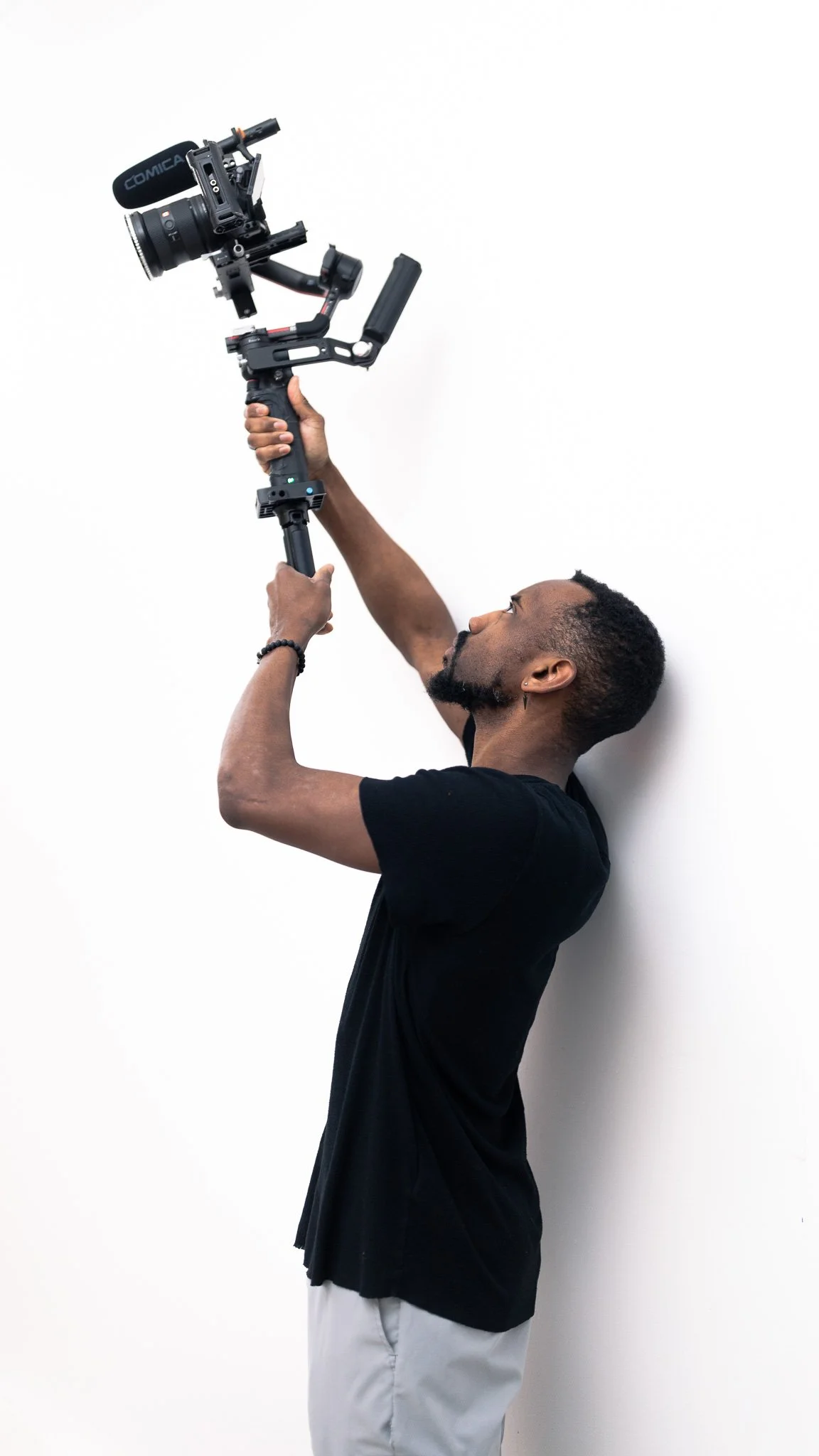 A man holding a professional camera mounted on a stabilizer against a plain white wall.