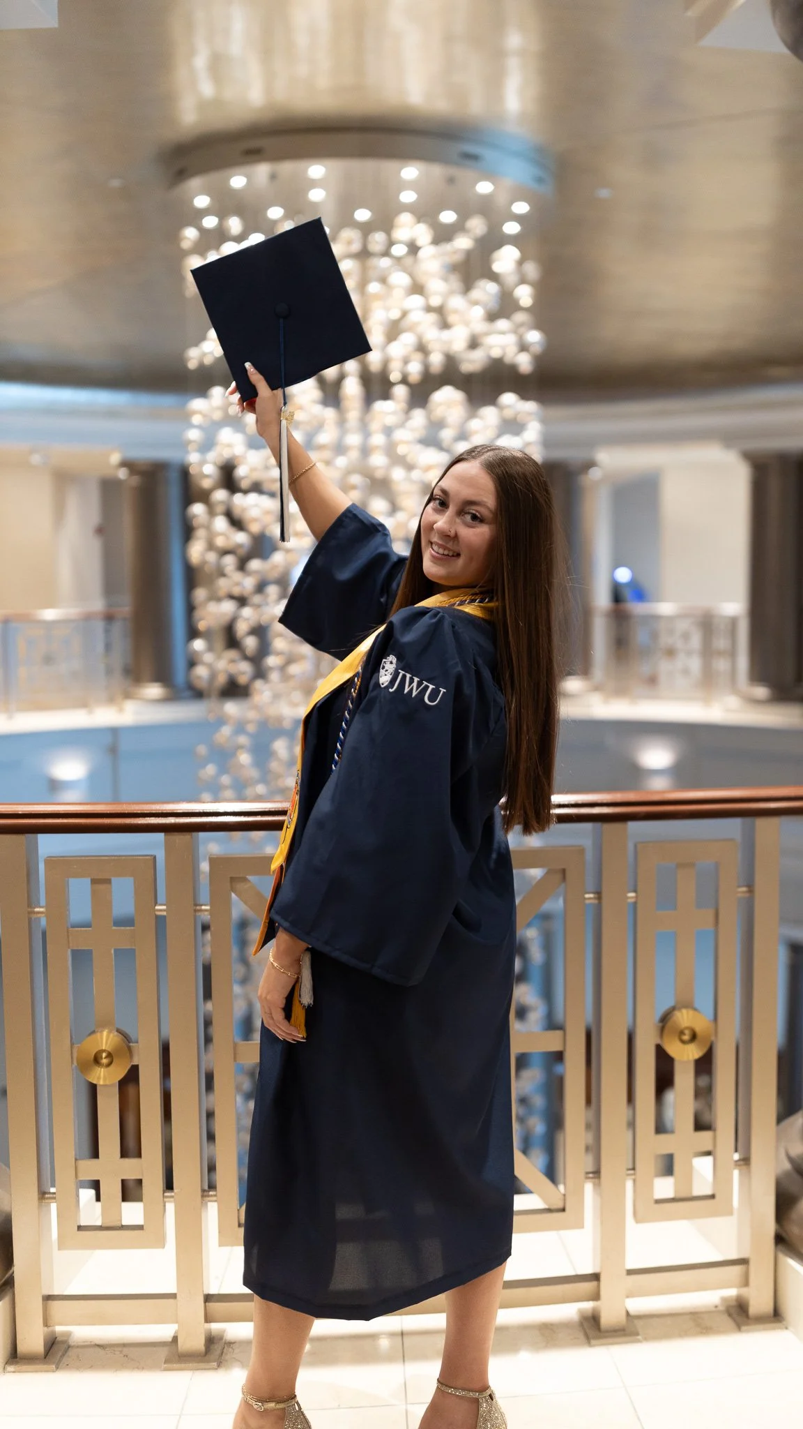 A woman in a graduation gown holding a graduation cap, smiling in an indoor setting with a chandelier and decorative railing in the background.