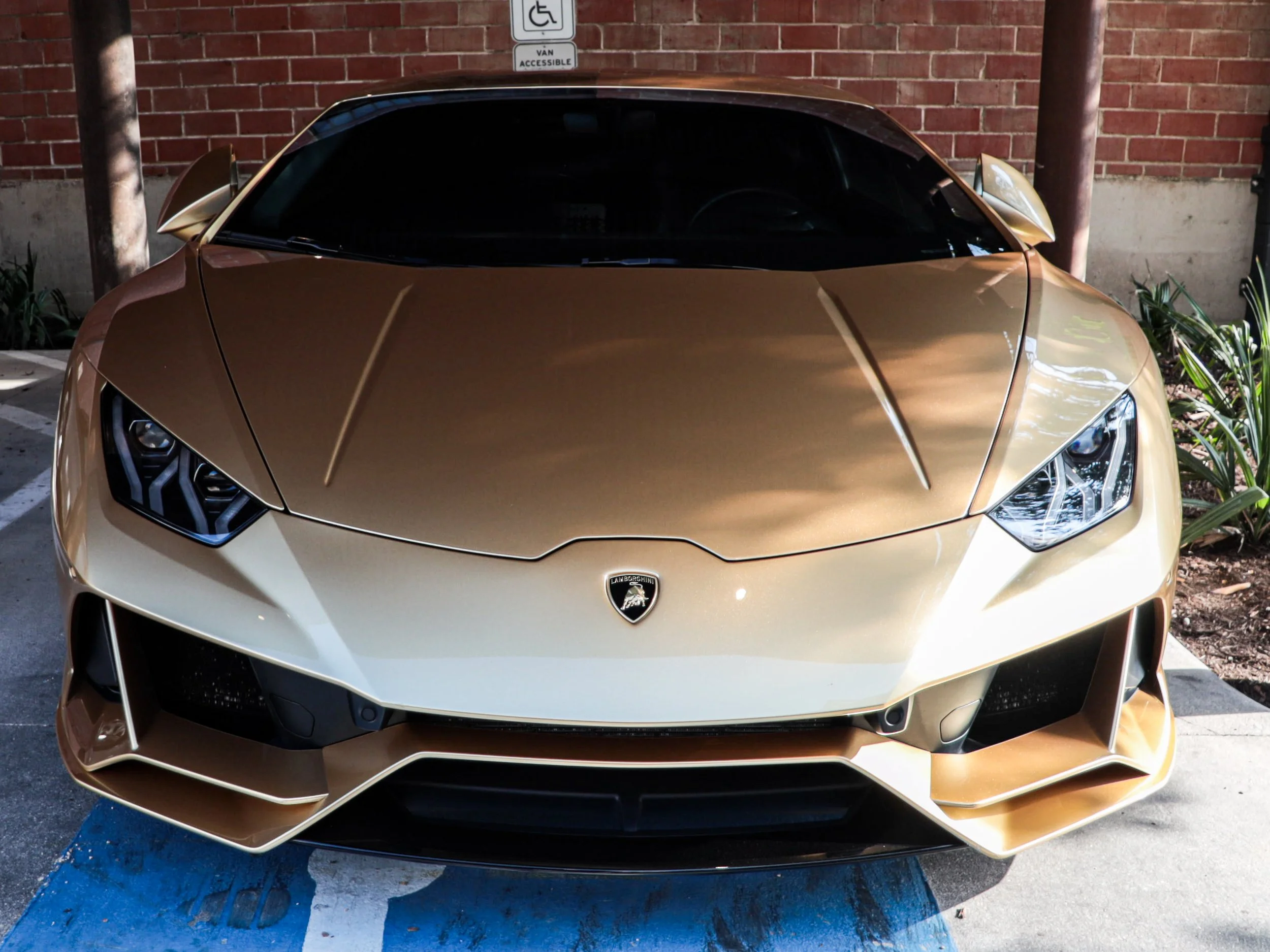 A gold-colored Lamborghini sports car parked in a parking space against a brick wall, with a sign indicating handicapped parking and vehicle accessibility.
