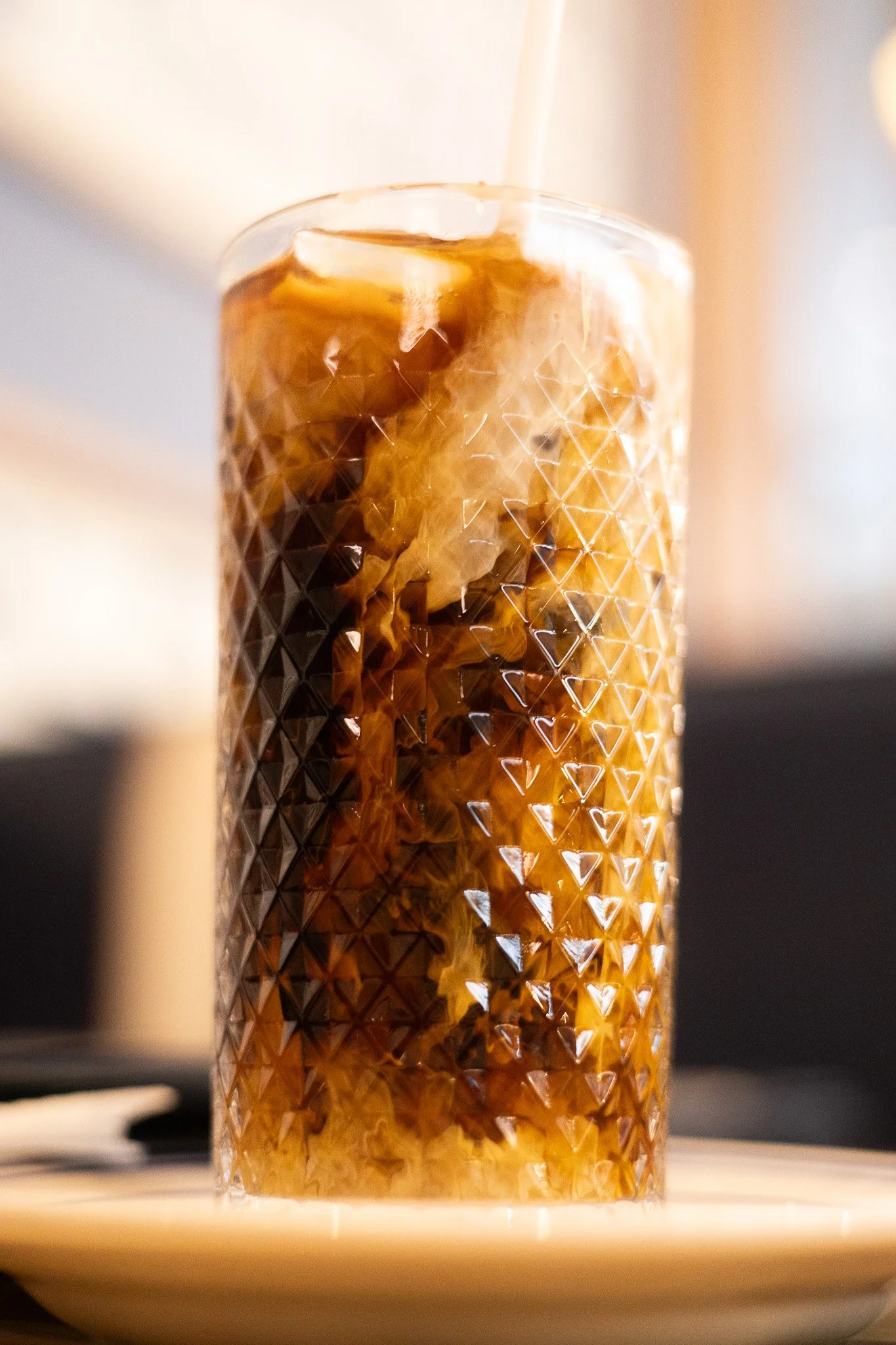 A glass with a diamond pattern contains a dark, fizzy beverage with ice and a slice of lemon, sitting on a saucer.