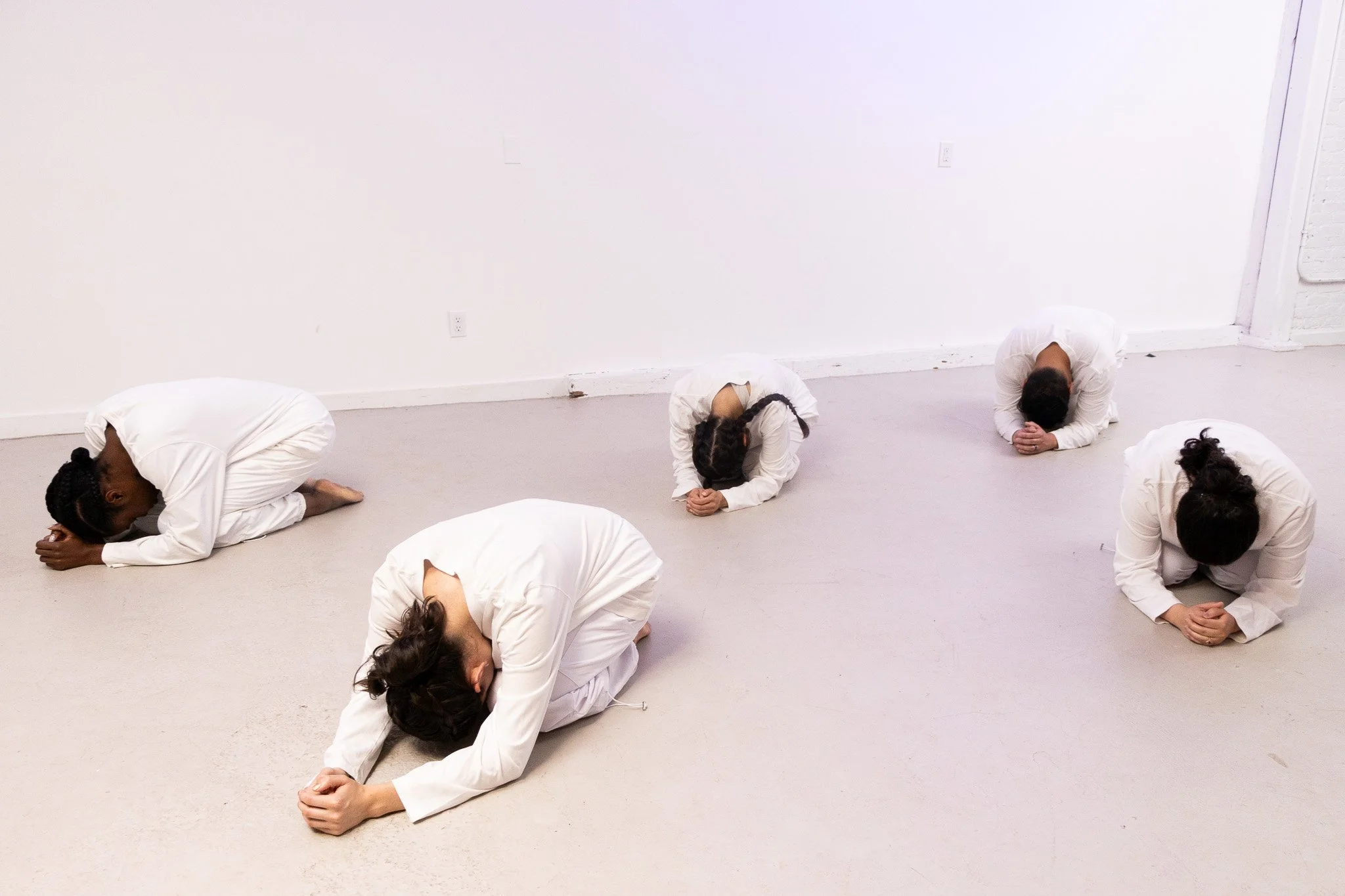 Multiple individuals dressed in white clothes practicing a yoga pose on a light-colored floor in a minimalist room with white walls.
