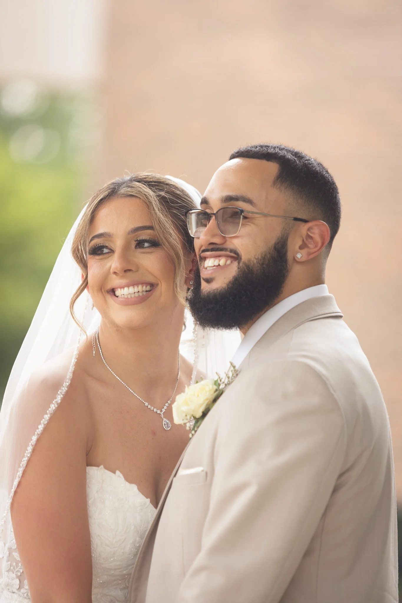A happy bride and groom smiling, dressed in wedding attire, standing outdoors with a blurred natural background.