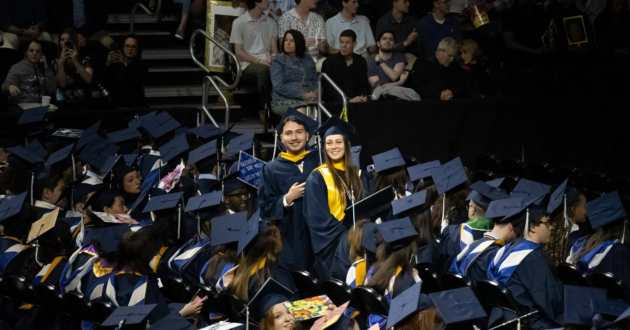 Graduates in caps and gowns celebrating at a graduation ceremony with audience seated behind them.